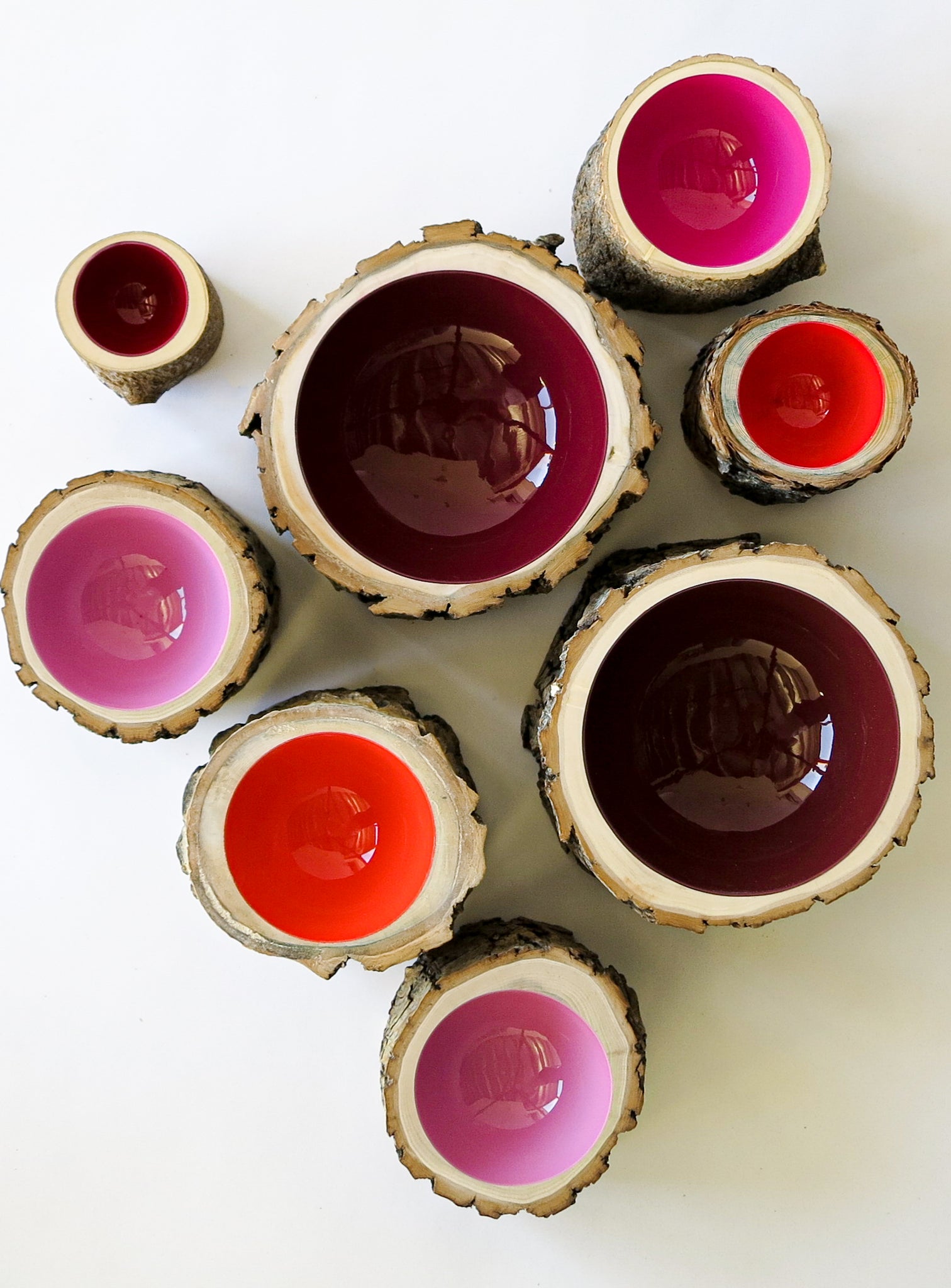 top view of a group of 8 Log Bowls in varying sizes and colours- 3 small wooden bowls with various barks, glossy interiors are bright red, dark current red, magenta and bright pink in colour.
